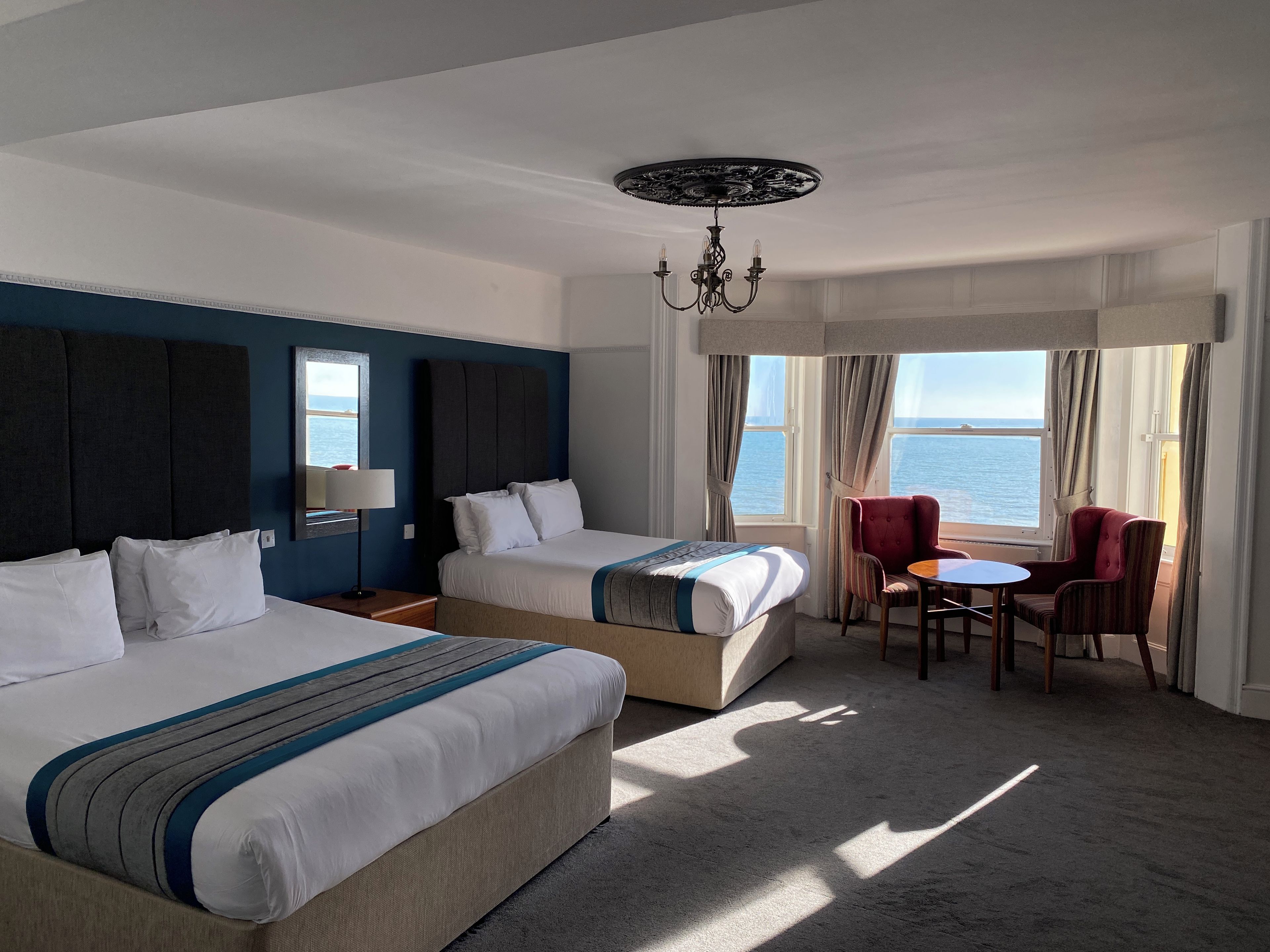 A hotel room with two beds, a dining table, and a view of the sea.