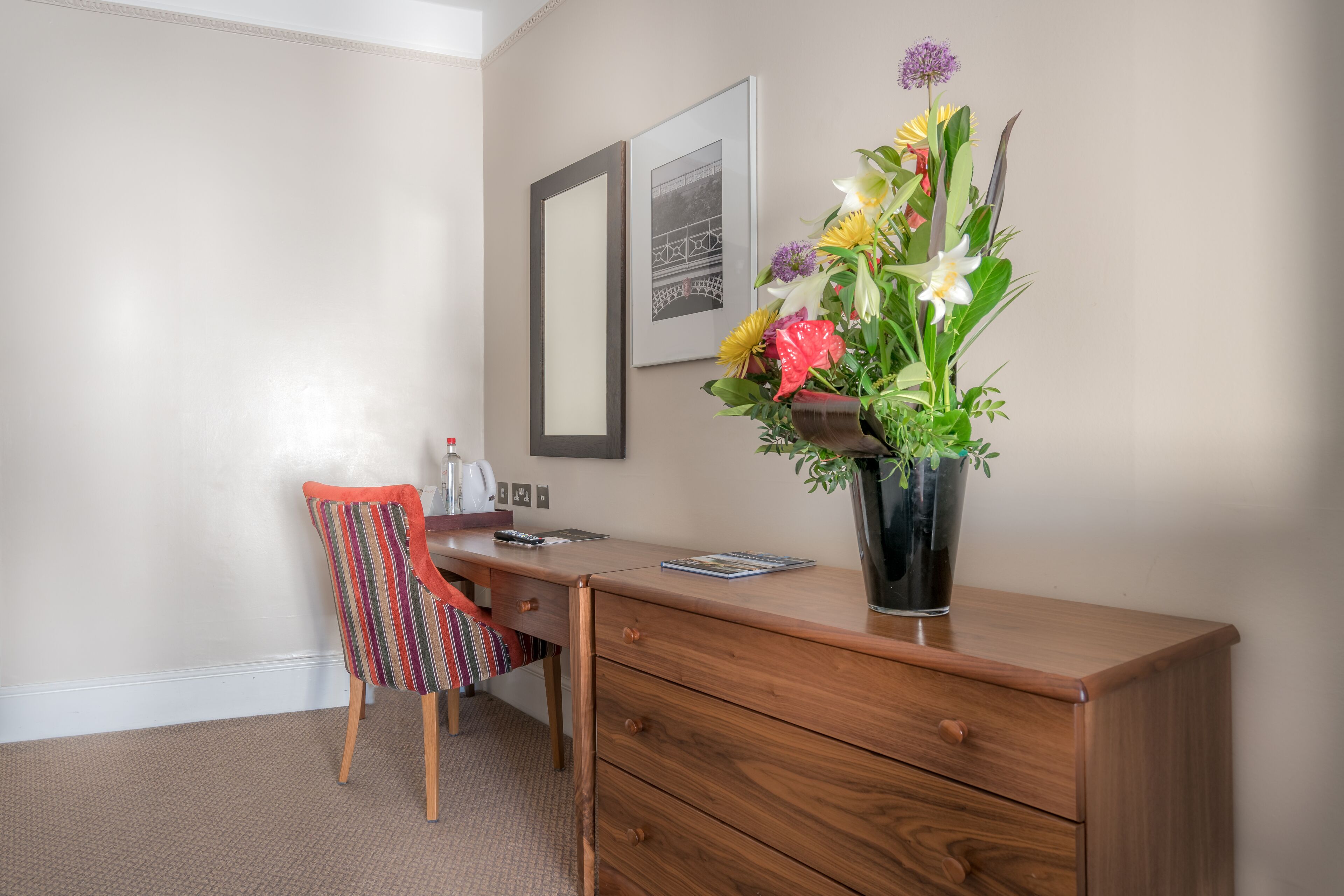 A wooden desk with a vase of flowers, a chair, and a framed picture on the wall.