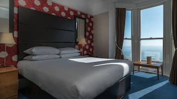 Habitación de hotel con una cama grande, una mesita de noche, una mesa y vistas al mar a través de un ventanal amplio.