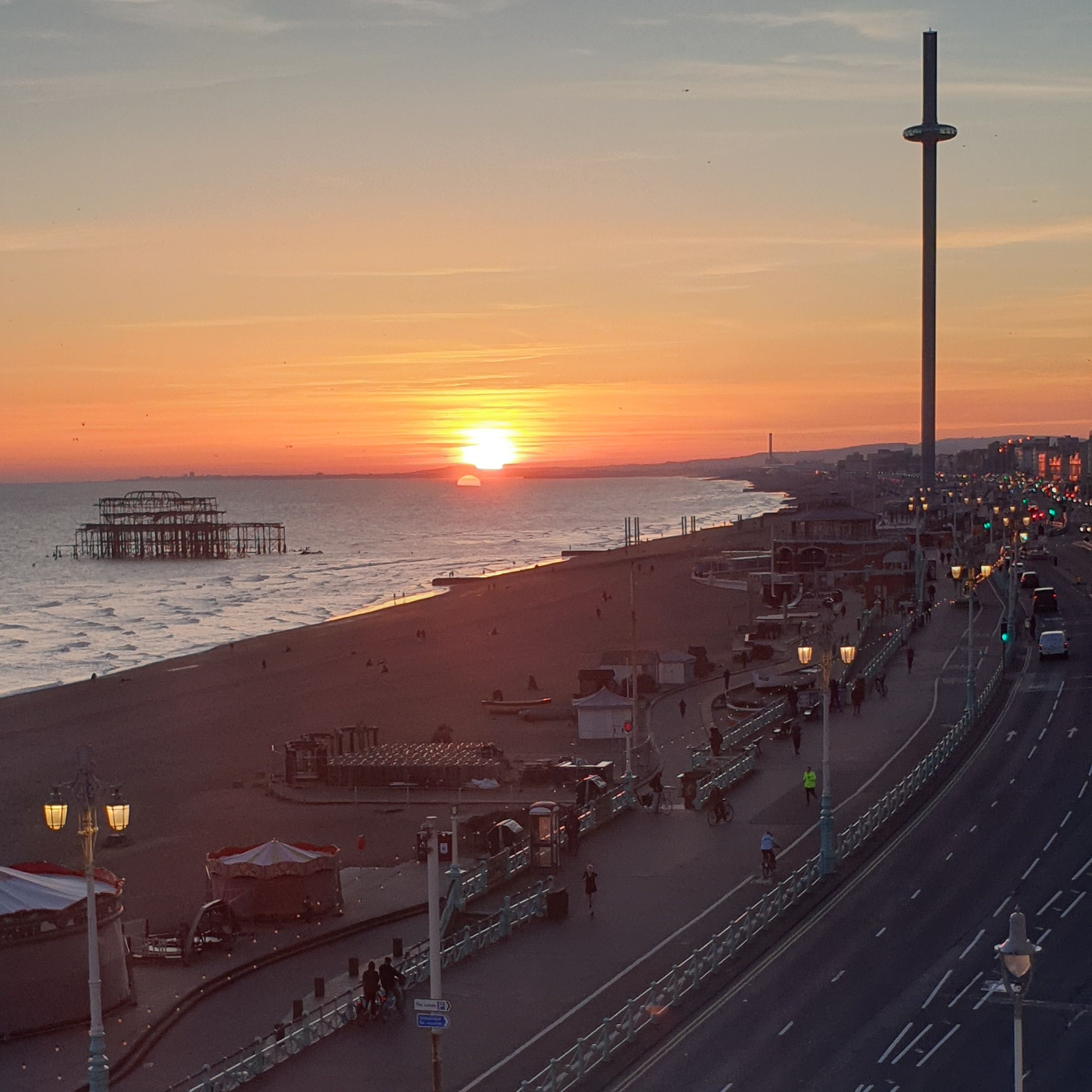 A coastal sunset view with a promenade, street lamps, and a pier.