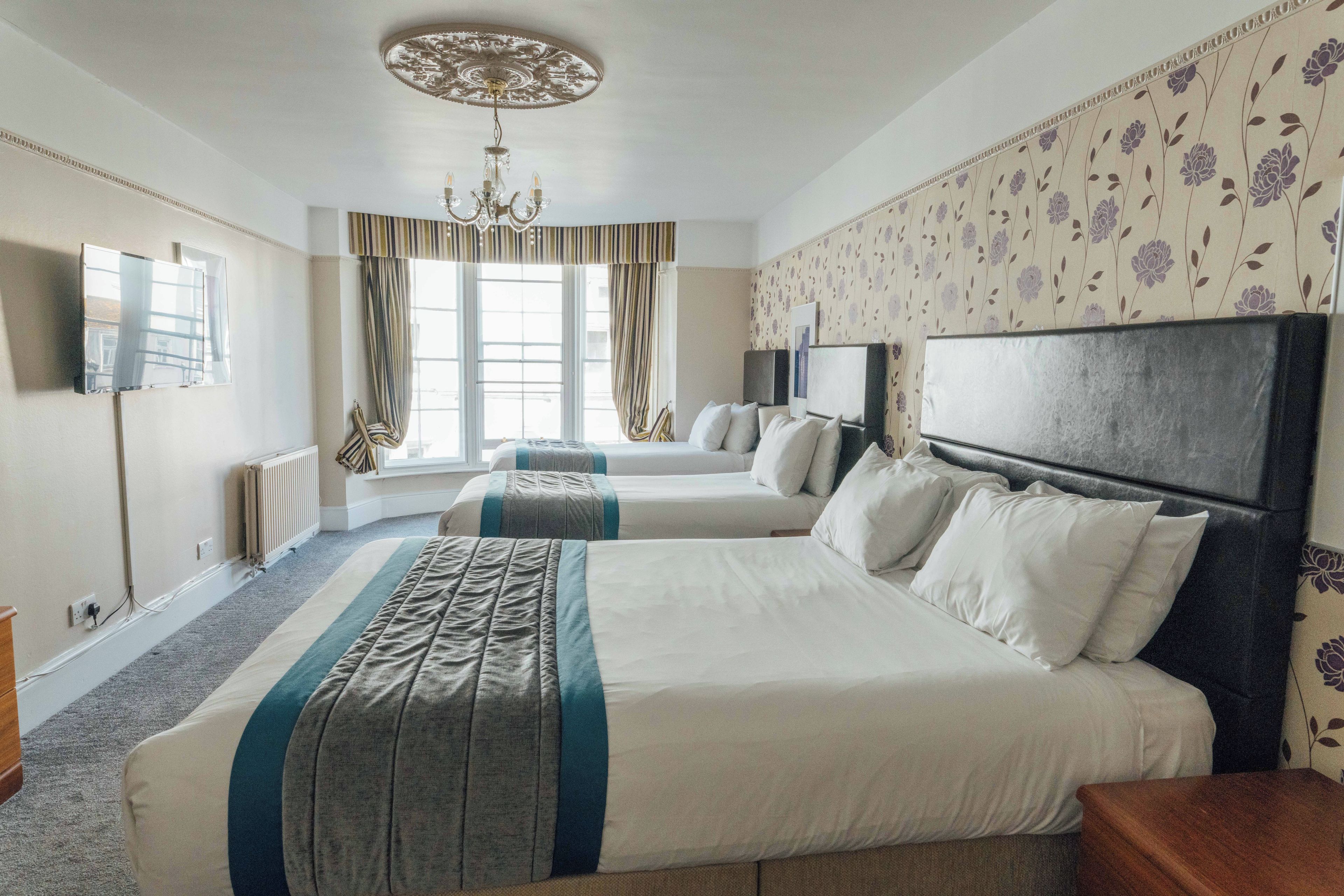 A hotel room with two beds, a large window with curtains, a flat-screen TV, and a chandelier.