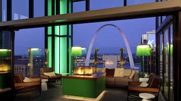 A modern lounge area with a central fire feature, seating, and large windows offering a city view.