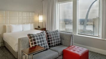 A hotel room with a bed, a sofa, a red ottoman, a nightstand, a lamp, and a view of a cityscape through large windows.