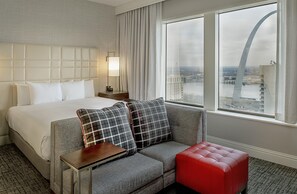 Habitación de hotel con cama, sofá, reposapiés rojo, mesita de noche, lámpara y vistas a un paisaje urbano a través de amplias ventanas.