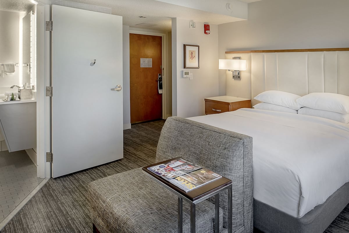 Habitación de hotel con cama, una mesita con revistas, mesita de noche, lámpara y puerta que da a otra habitación.