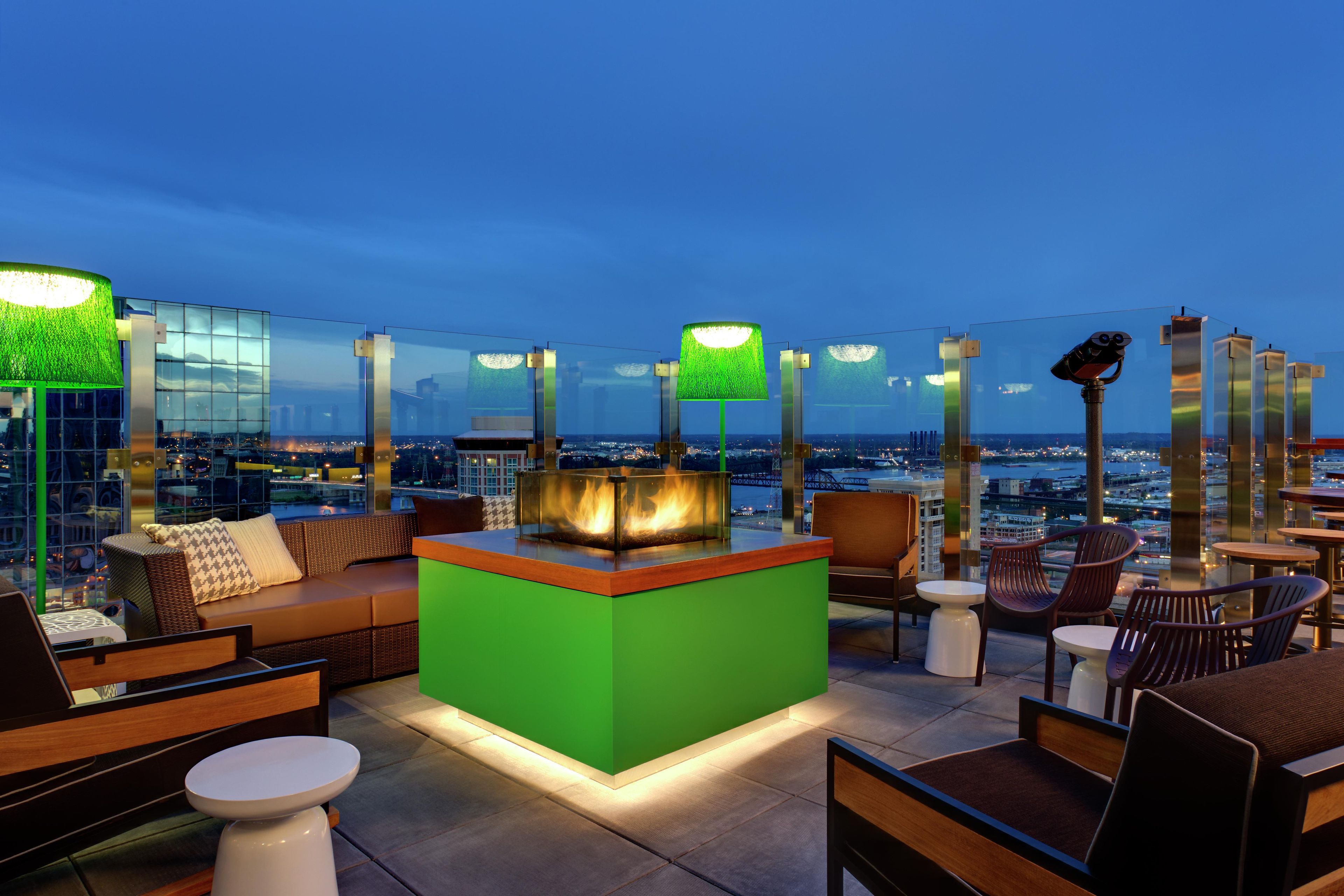 An outdoor seating area with a green bar, lounge chairs, and a view of the cityscape at dusk.