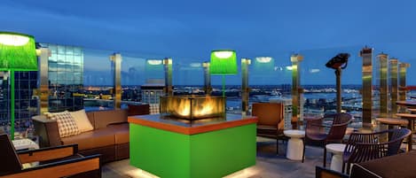 An outdoor seating area with a green bar, lounge chairs, and a view of the cityscape at dusk.