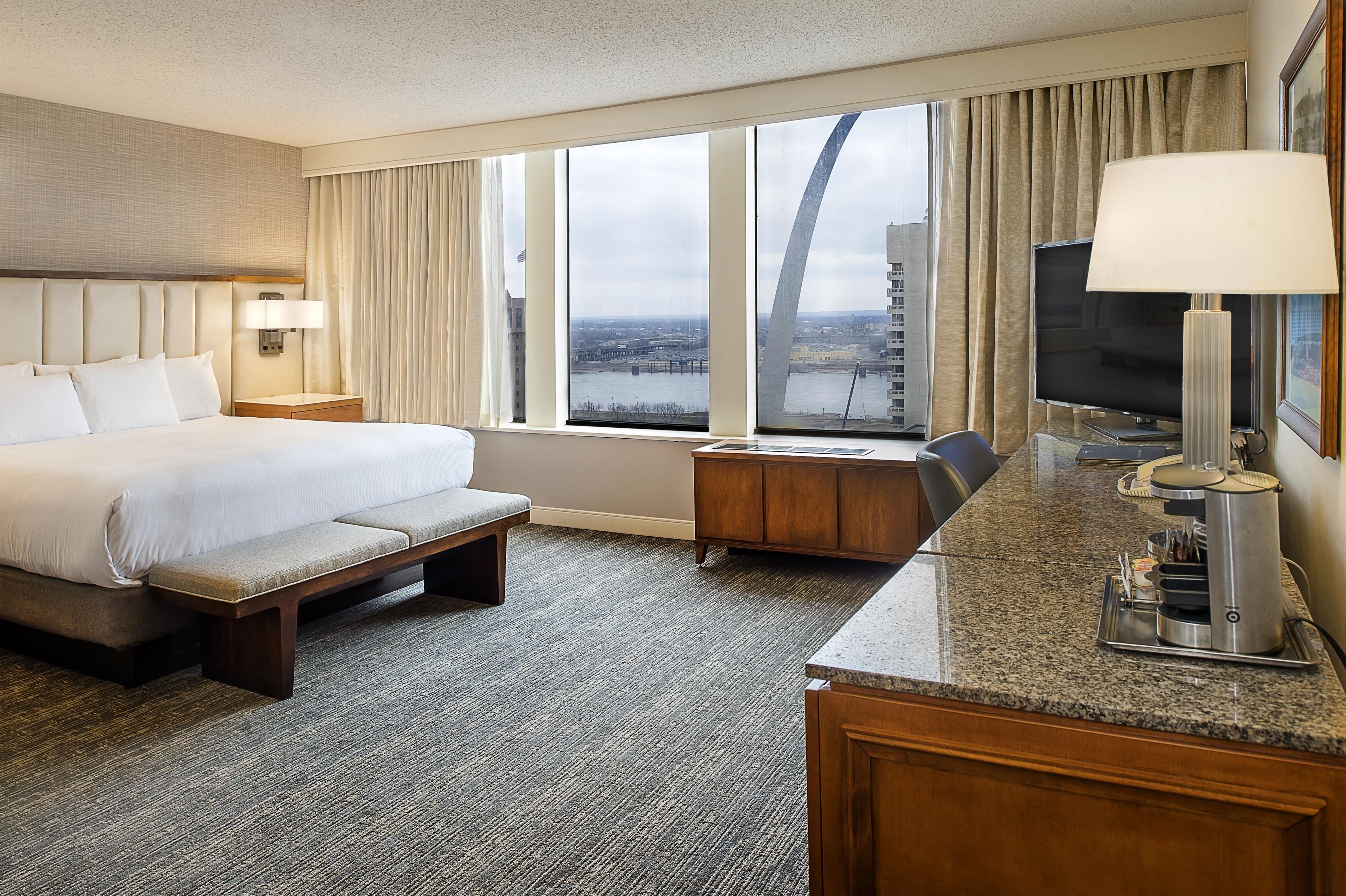 A hotel room with a bed, a desk with a lamp, a TV, and a view of a bridge and cityscape.