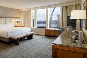 A hotel room with a bed, a desk with a lamp, a TV, and a view of a bridge and cityscape.