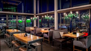 Un espace bar moderne avec de hautes tables et des tabourets, de grandes fenêtres offrant une vue sur la ville et des lustres verts.