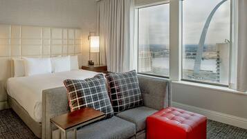 A hotel room with a large bed, a gray sofa with plaid cushions, a red ottoman, and a view of a cityscape through a large window.