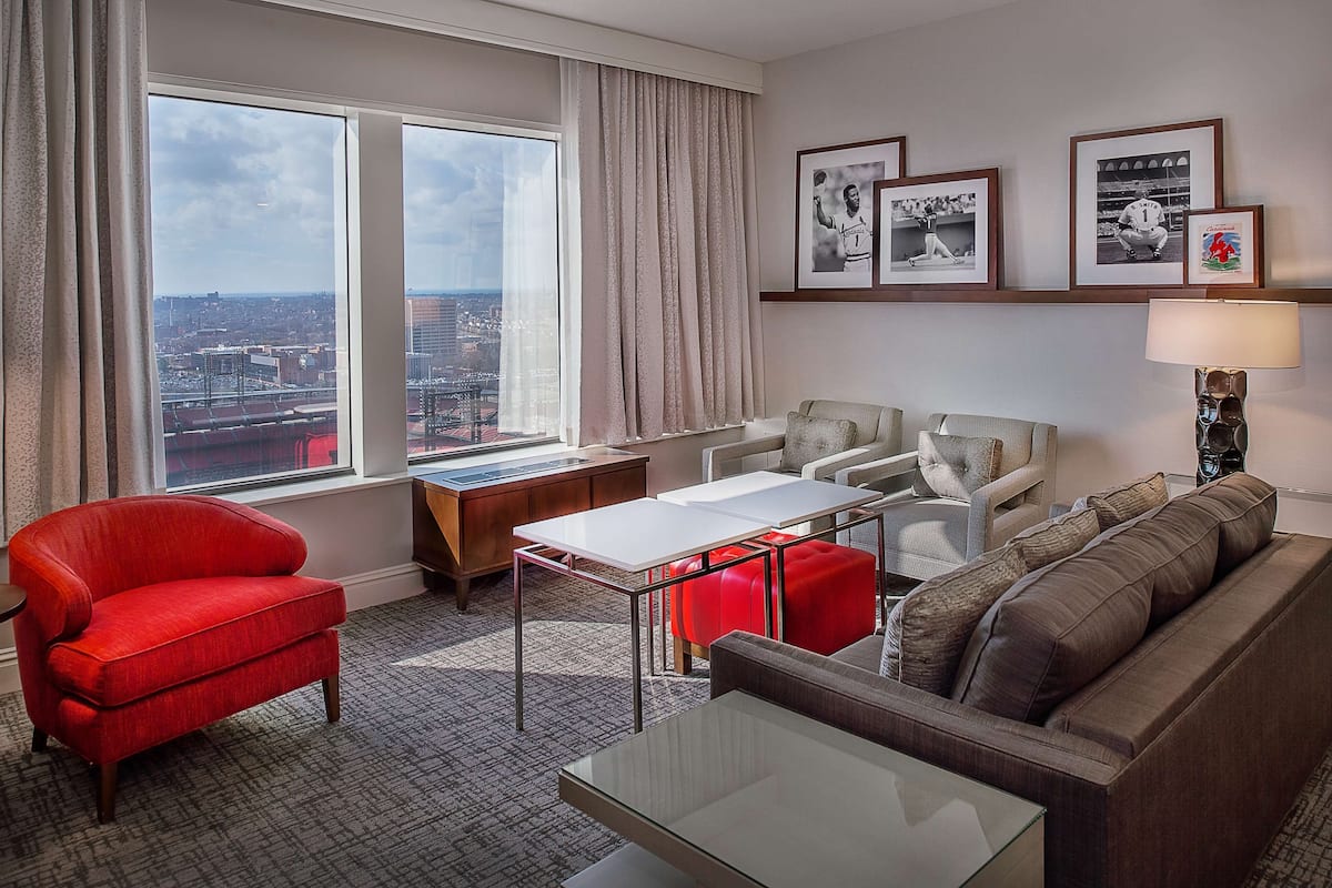 Habitación de hotel con vistas a la ciudad, un sillón rojo, un sofá gris, una mesita de café blanca y recuerdos deportivos enmarcados en la pared.