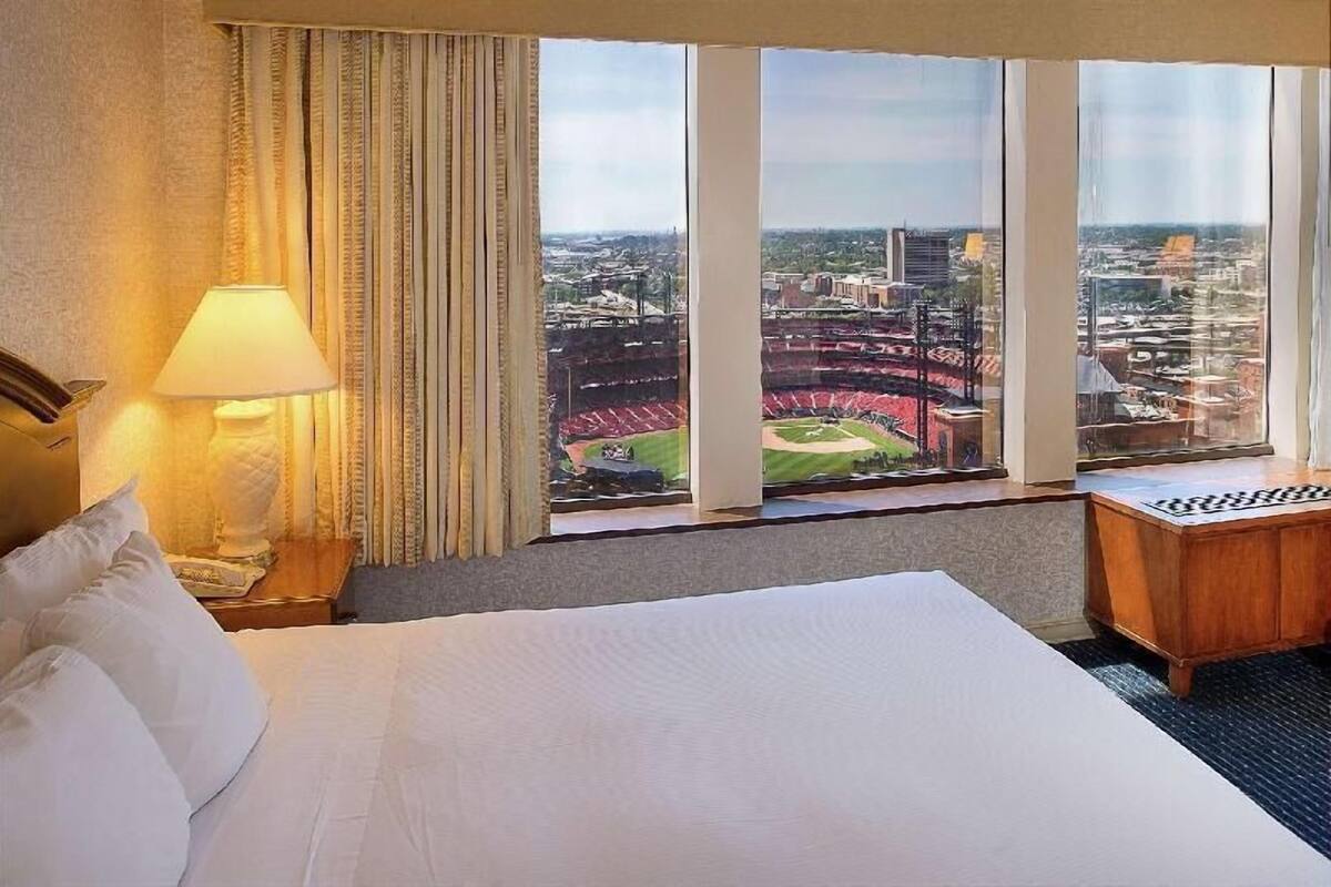Habitación de hotel con una cama, una lámpara de noche, una mesita de noche de madera y vistas a un estadio a través de grandes ventanales.