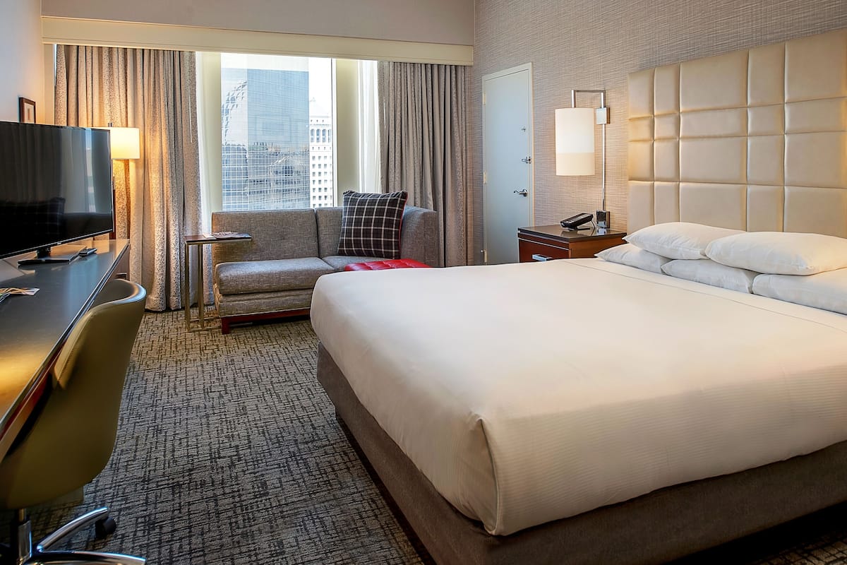 Una habitación de hotel con una cama grande, un escritorio con televisor, una silla, una mesita de noche y una ventana con cortinas.