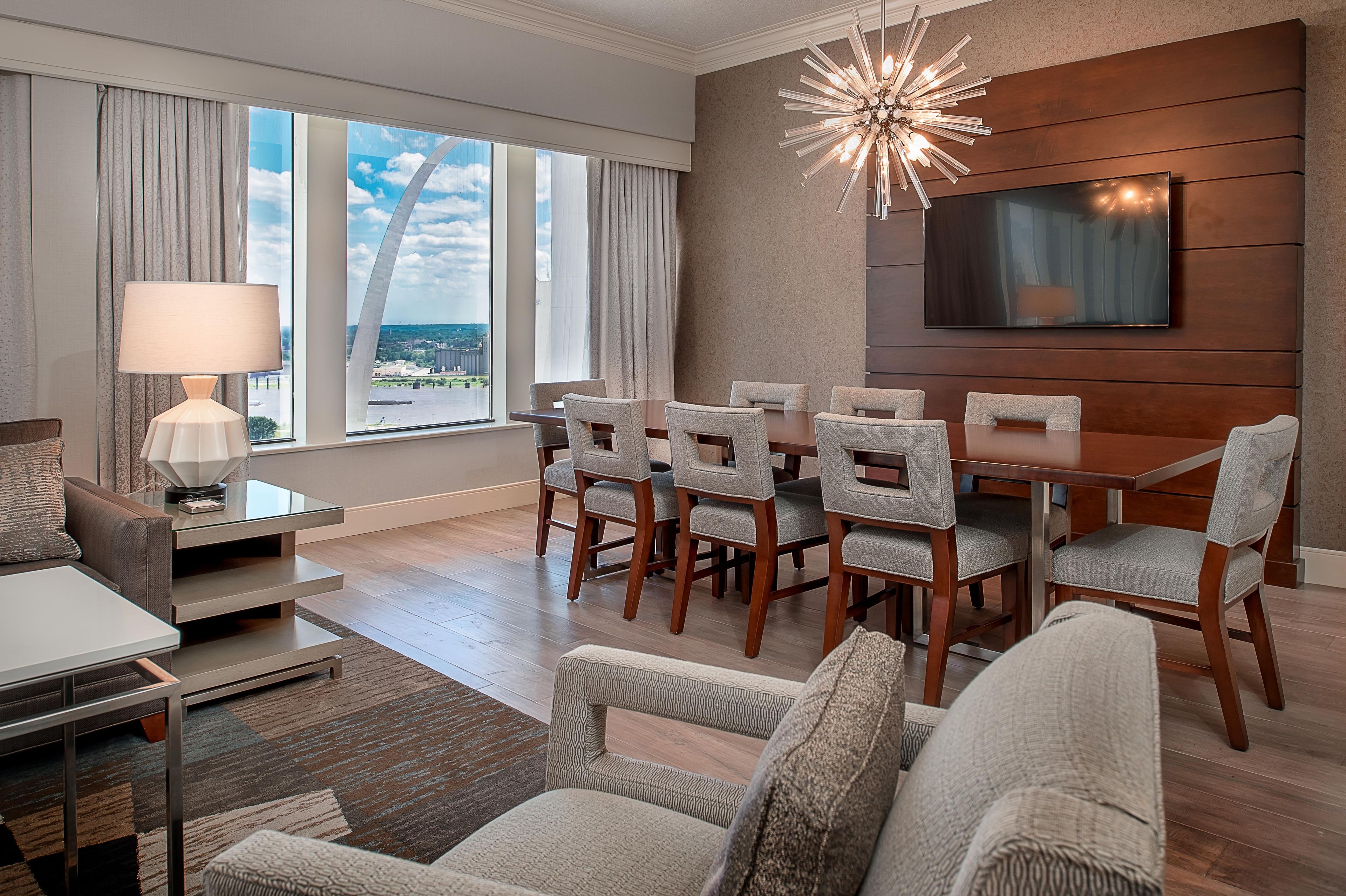 A modern hotel room with a dining area, a sofa, a TV mounted on the wall, and large windows with curtains.