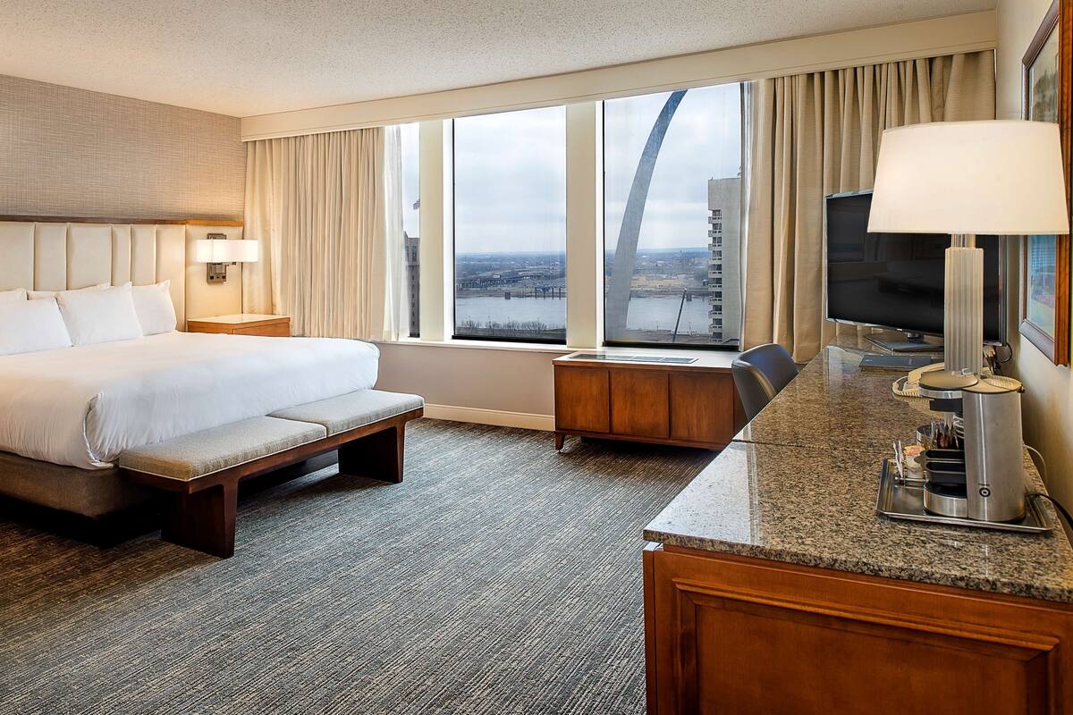 Habitación de hotel con una cama grande, un escritorio con televisor, cafetera y vistas a un puente y al paisaje urbano.