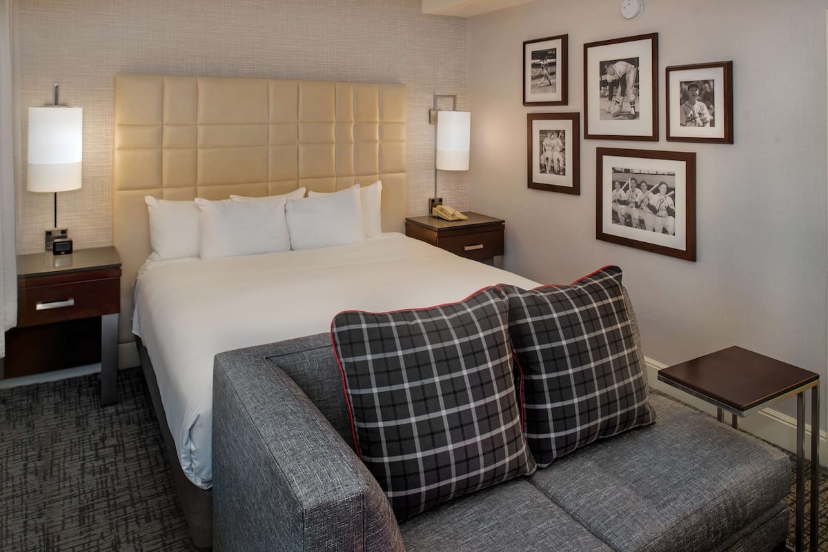 Una habitación de hotel con una cama grande, dos mesitas de noche, una pequeña mesa y un sofá. Hay cuadros en la pared.