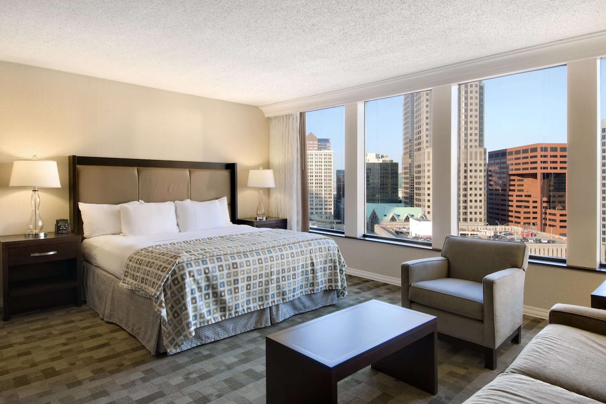 Habitación de hotel con una cama grande, dos mesitas de noche con lámparas, una silla, una mesa de centro y vistas a edificios urbanos a través de grandes ventanales.