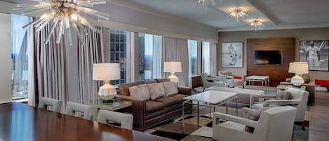 A modern hotel room with a large dining table, comfortable seating, a sofa, and a television.