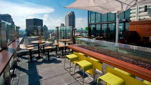 Un bar situé sur le toit, doté d’une rambarde en verre, de tabourets de bar jaunes et d’un grand parasol.