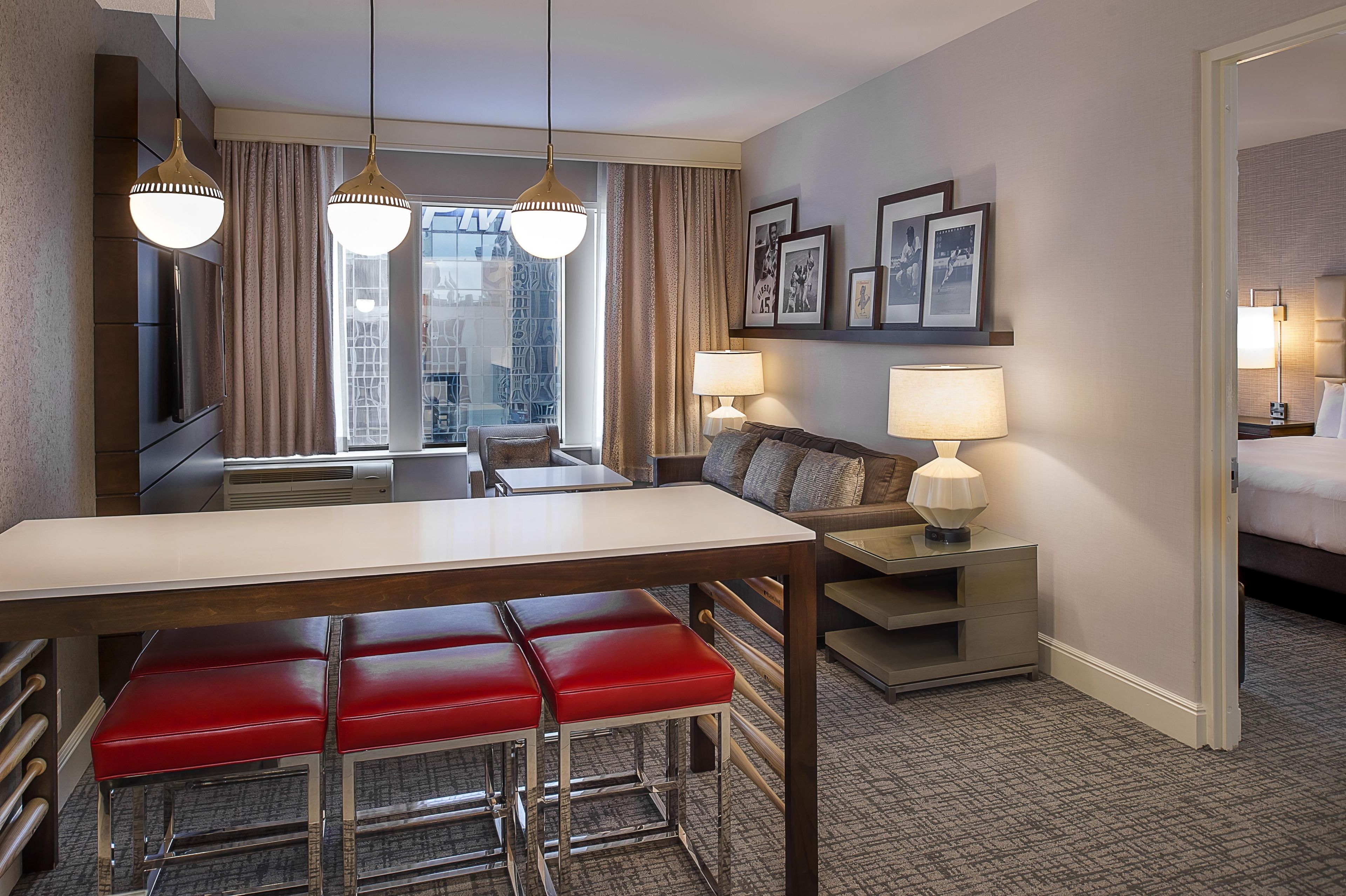 A hotel room with a dining area, a sofa, a bed, and a view of the cityscape through the window.