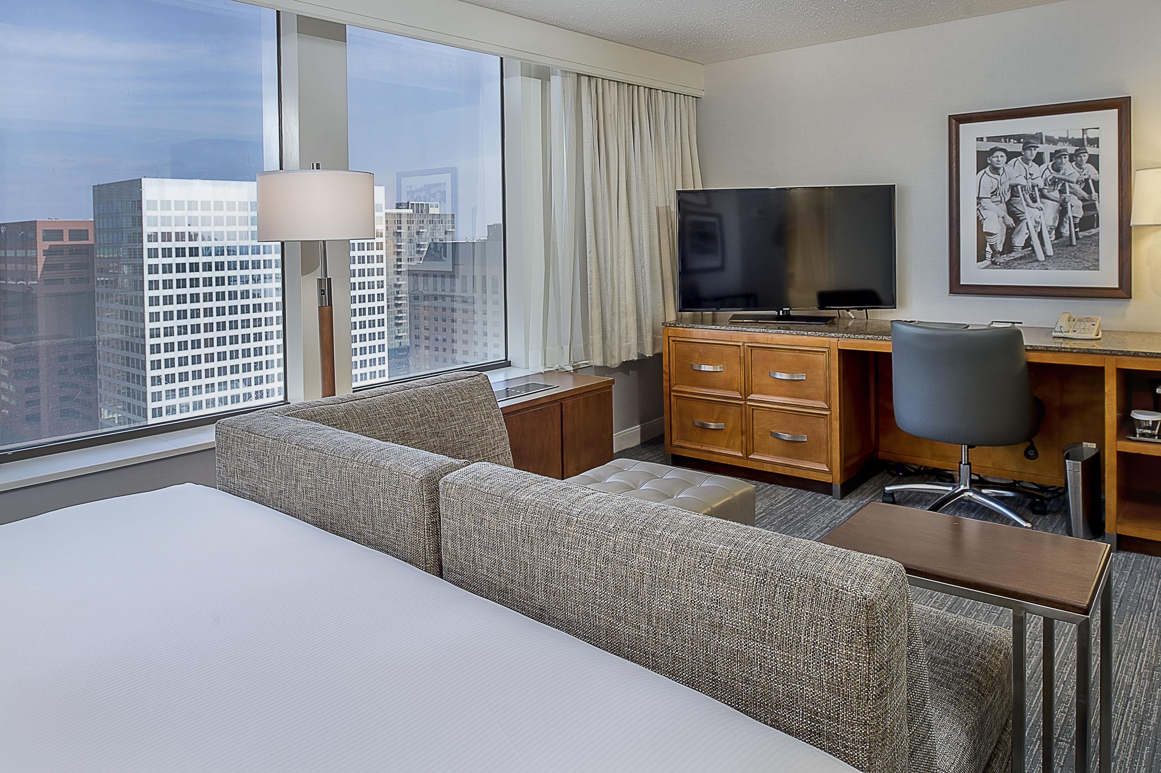 Habitación de hotel con una ventana grande, una cama, un escritorio con televisor y un cuadro en la pared.