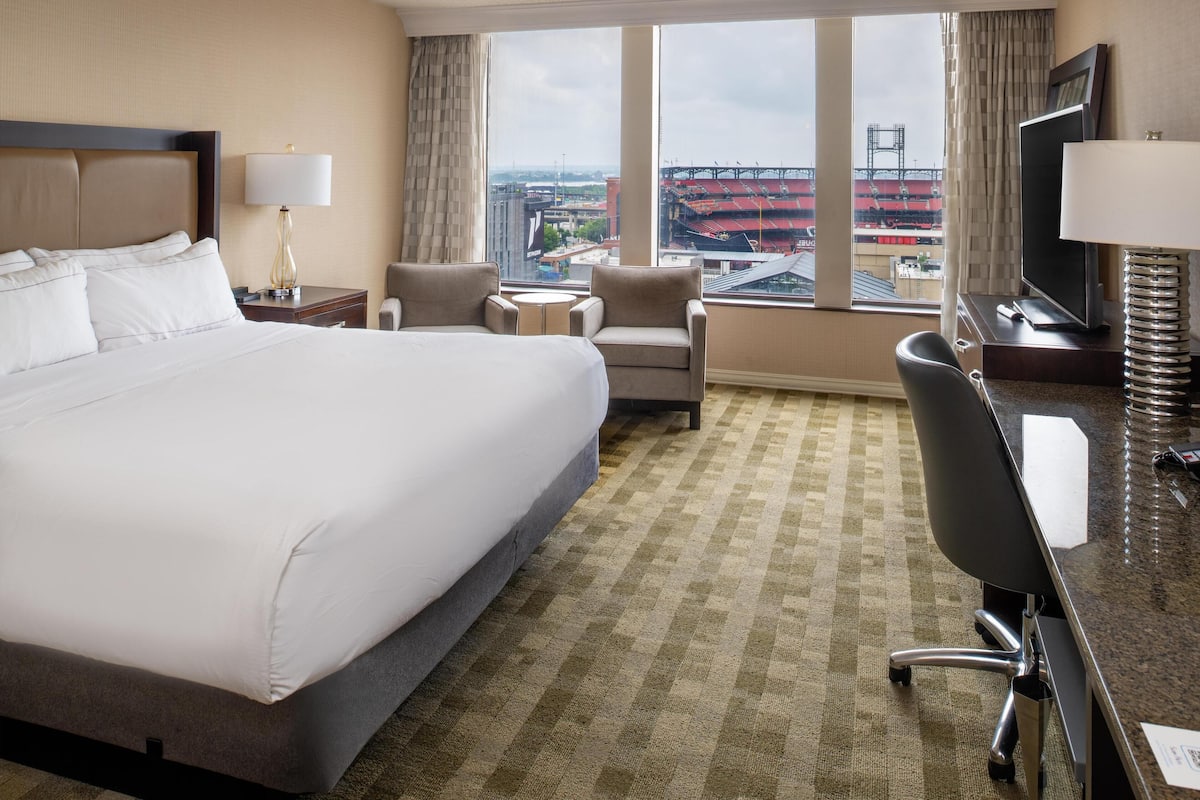 Habitación de hotel con una cama grande, dos sillones, un escritorio con televisor y vistas a un estadio desde la ventana.