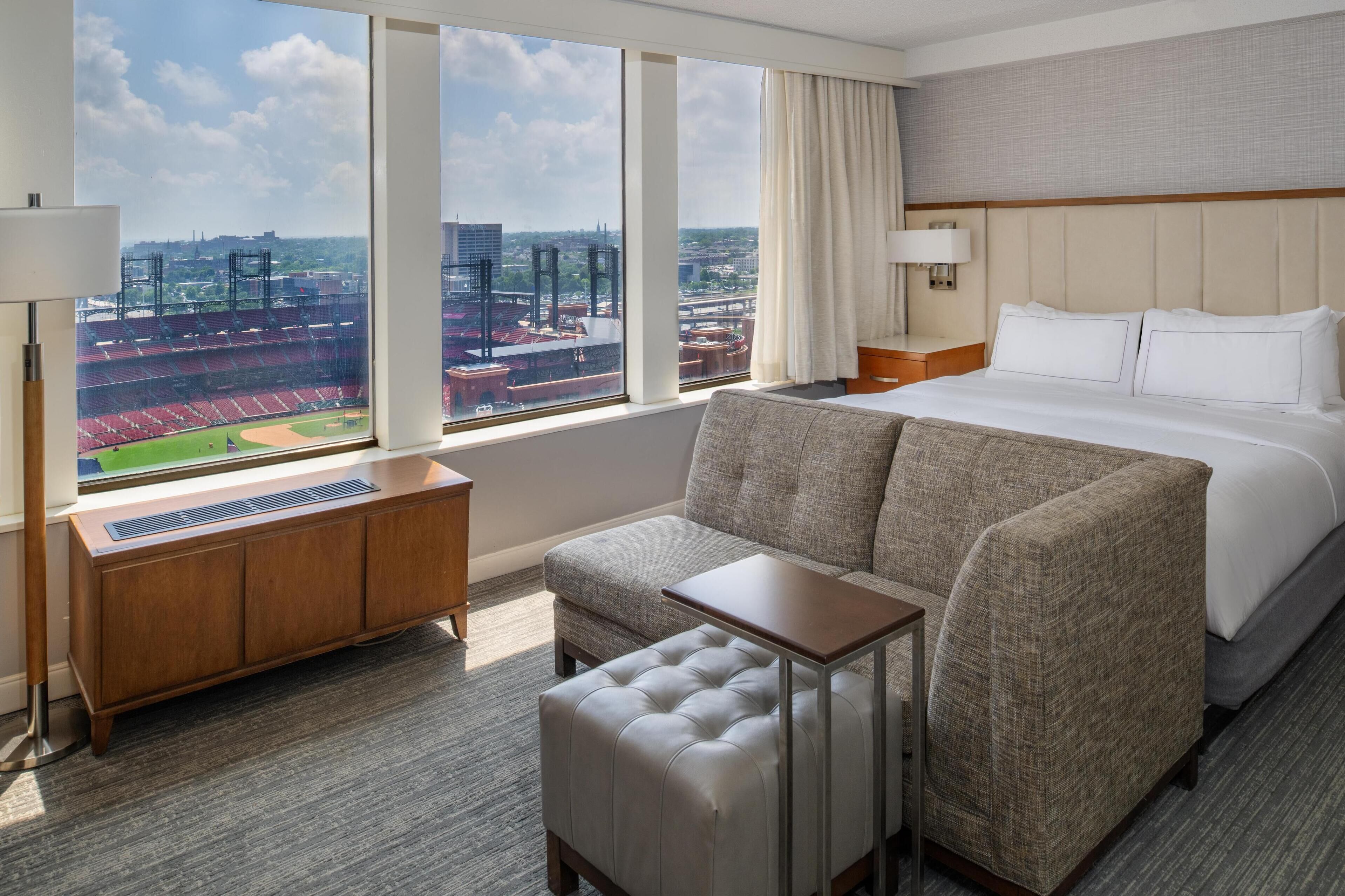 Habitación de hotel con un ventanal grande que ofrece vistas a un estadio y al paisaje urbano. La habitación tiene una cama, un sofá, una mesita y un armario de madera.