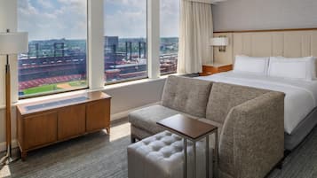 Habitación de hotel con un ventanal grande que ofrece vistas a un estadio y al paisaje urbano. La habitación tiene una cama, un sofá, una mesita y un armario de madera.