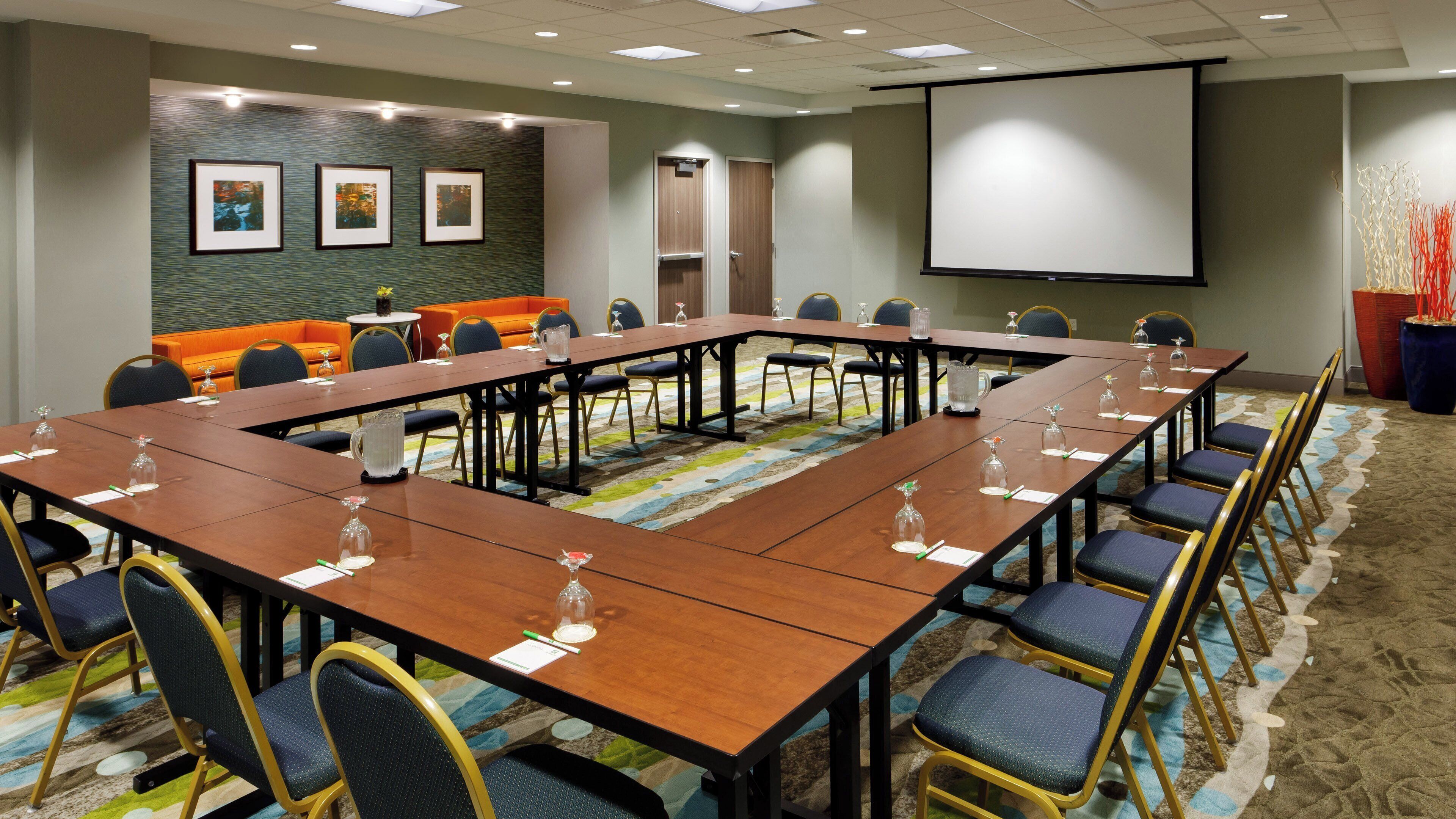 A conference room with a long table, chairs, a projector screen, and a seating area with orange chairs.