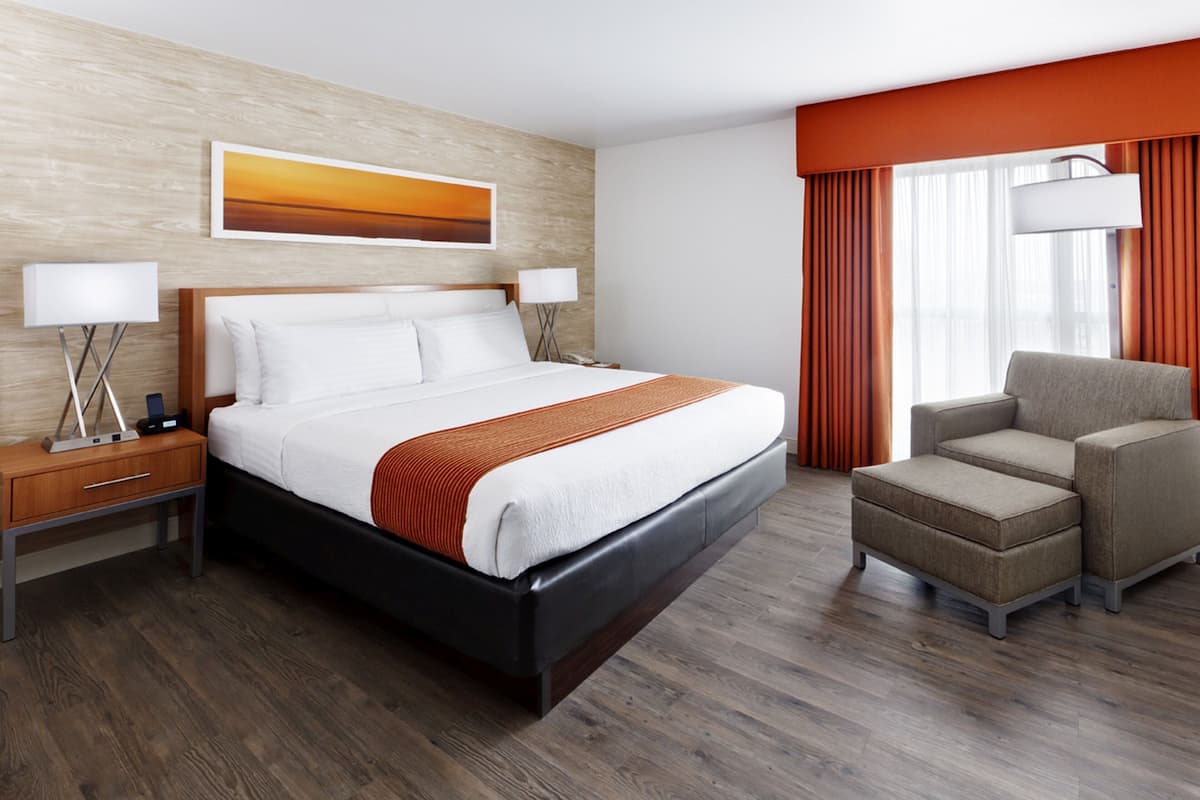 Una habitación de hotel con una cama grande, una mesita de noche, una silla y un ventanal con cortinas.