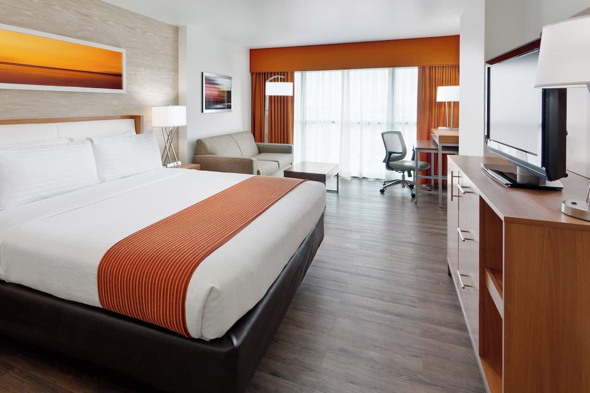 Habitación de hotel con una cama grande, un escritorio con silla, televisión y ventana con cortinas.