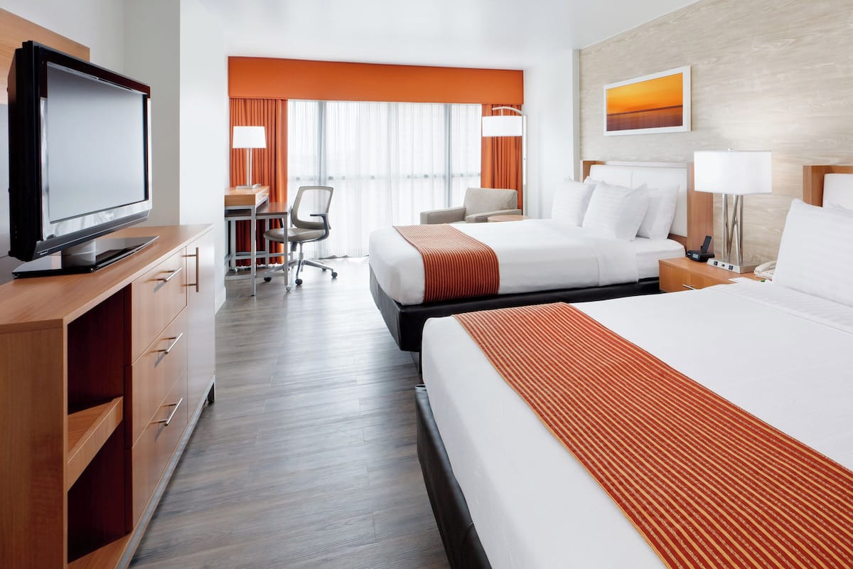 Habitación de hotel con dos camas, un escritorio, una silla, un televisor y un ventanal con cortinas.