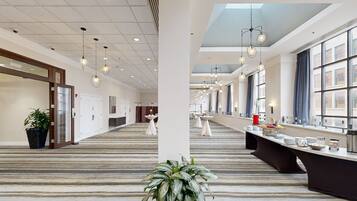 A hotel lobby with a long reception desk, multiple tables, and a large window with curtains.