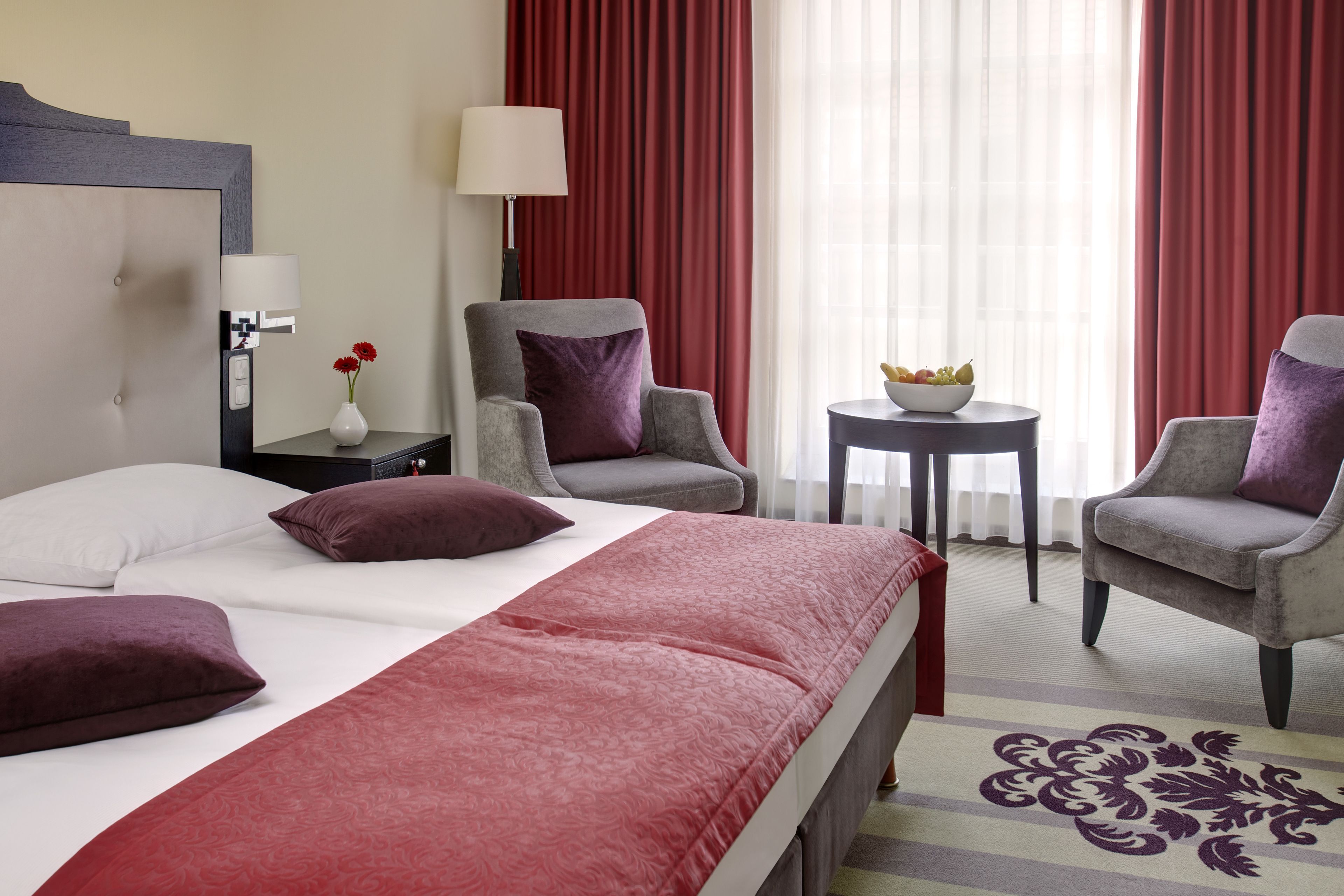 A hotel room with a bed, two armchairs, a small table with a bowl of fruit, a lamp, and a vase with flowers.