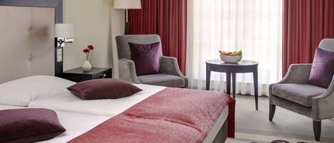 A hotel room with a bed, two armchairs, a small table with a bowl of fruit, a lamp, and a vase with flowers.