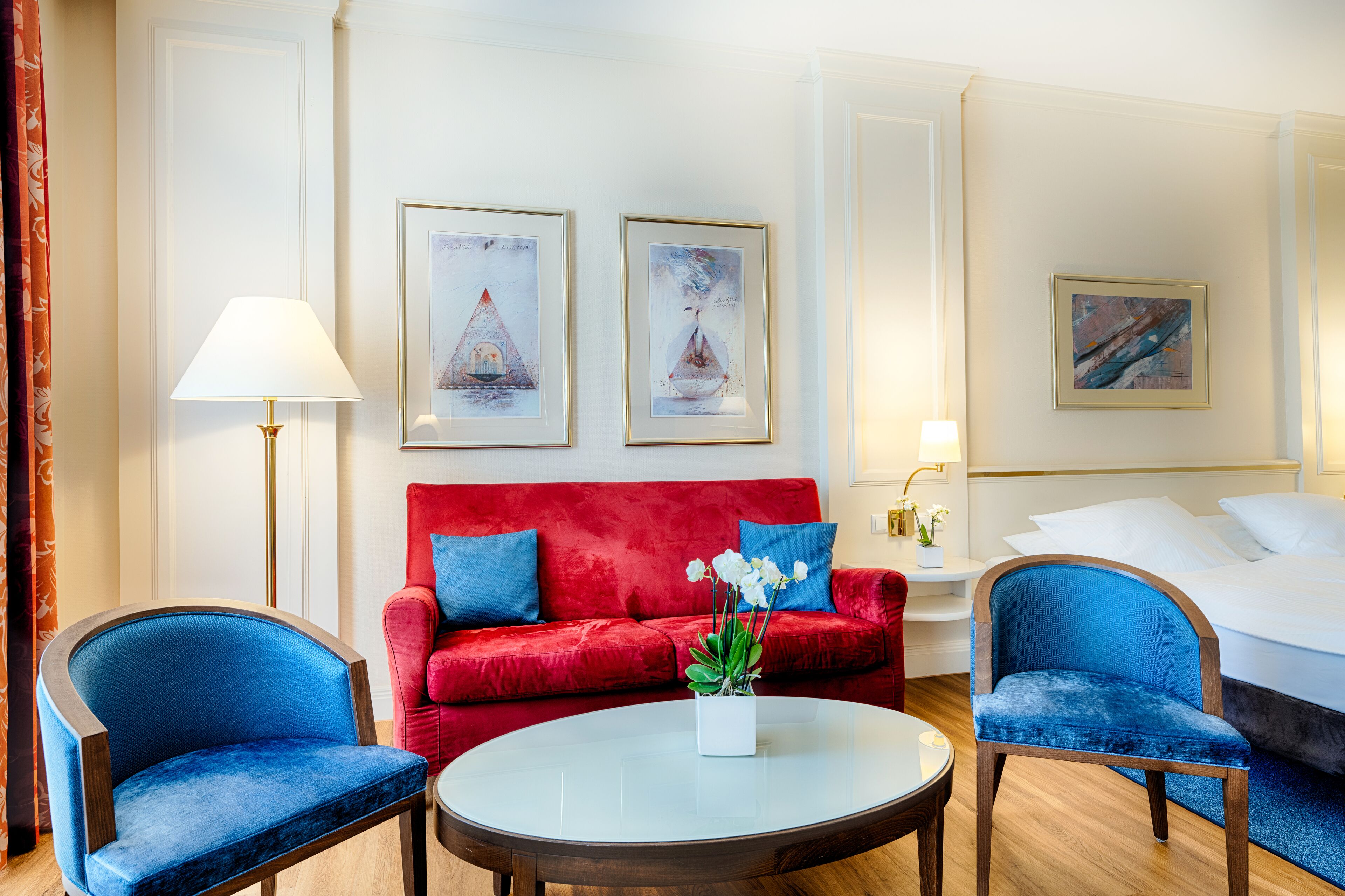 Ein Hotelzimmer mit einem roten Sofa, zwei blauen Sesseln, einem runden Couchtisch mit weißer Vase und einem Bett mit weißer Bettwäsche.