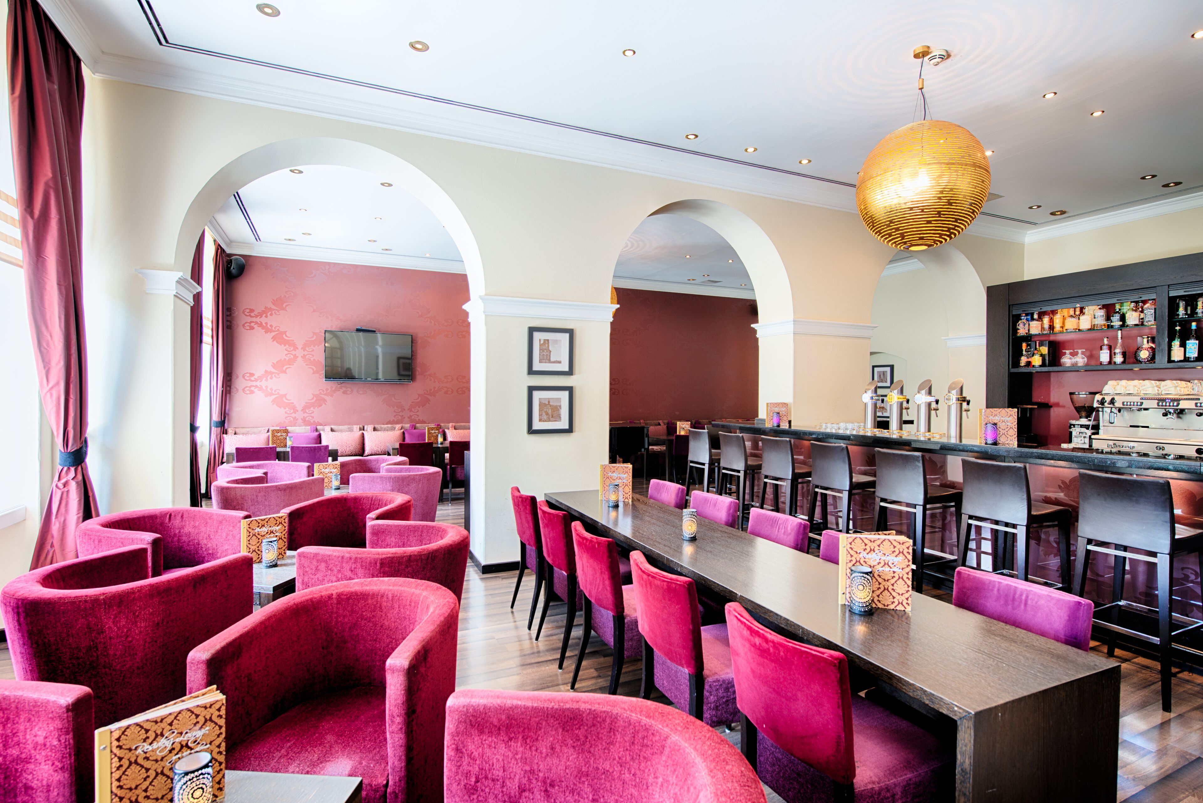 A restaurant with red upholstered chairs, a long wooden table, and a bar area with shelves of bottles.