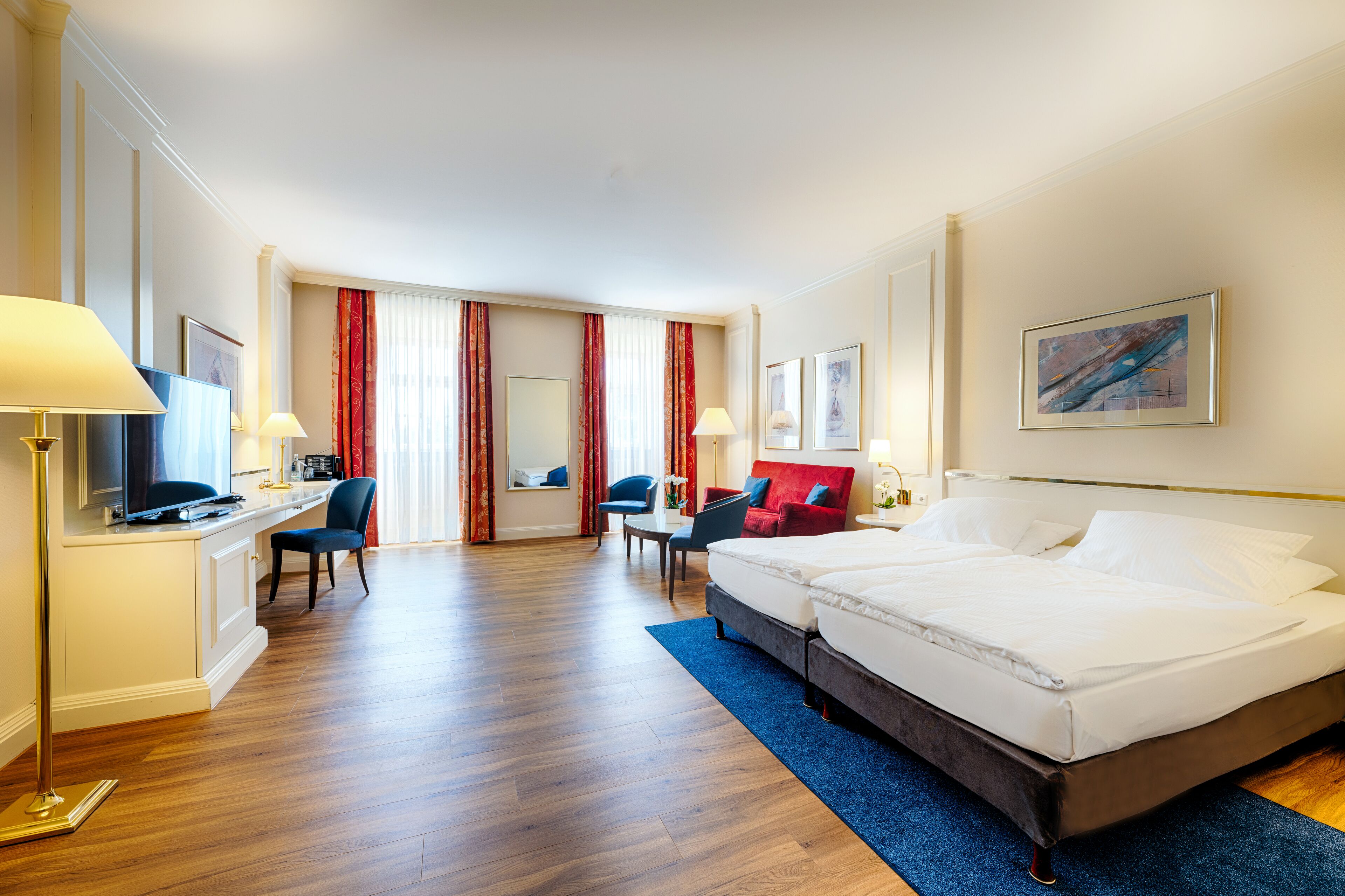 Una habitación de hotel con cama, dos sillas, escritorio, televisor y ventana con cortinas.