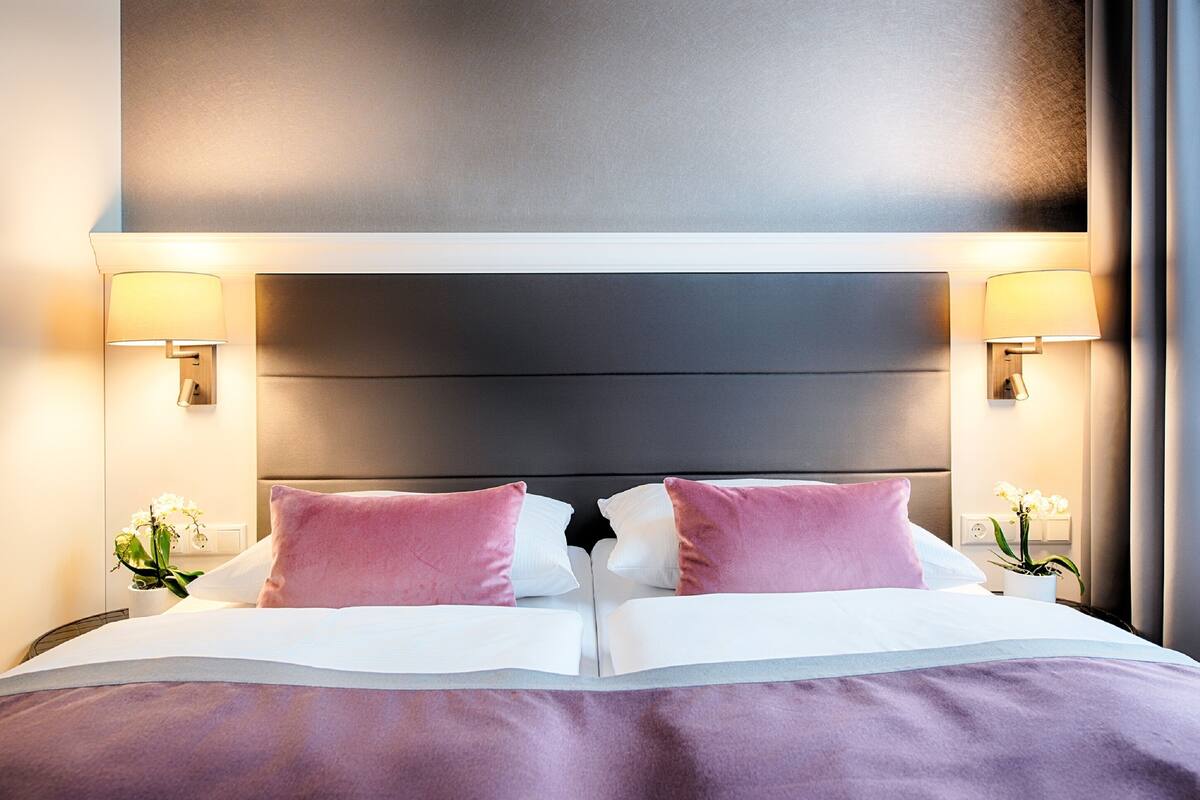 Ein ordentlich bezogenes Bett mit weißer und rosa Bettwäsche, zwei Nachttischlampen und ein kleiner Topf mit einer Orchidee auf dem Nachttisch.