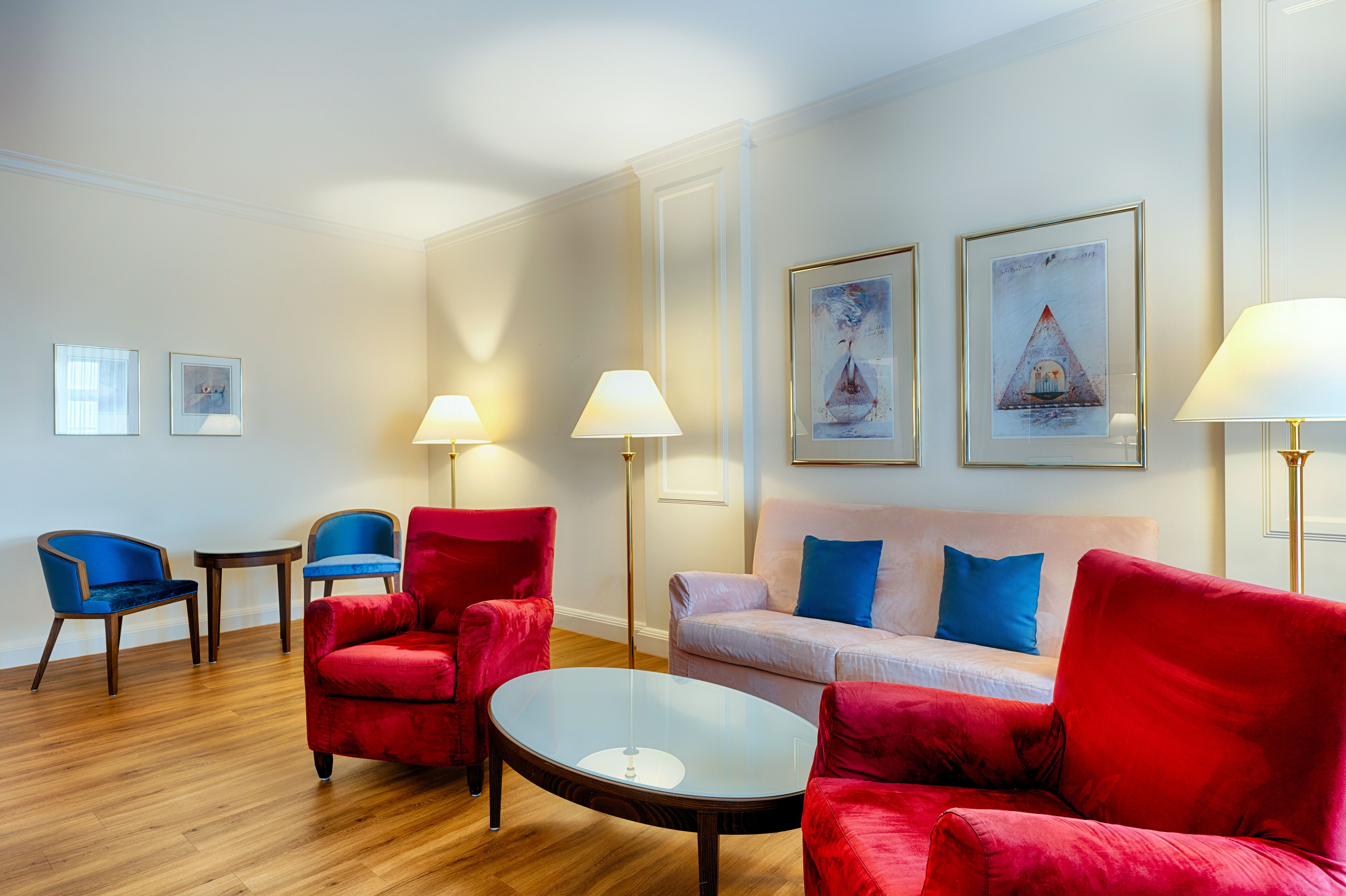 Uma sala de estar moderna com piso de madeira, cadeiras vermelhas e azuis, uma mesa de centro redonda de vidro, um sofá de cor clara com almofadas azuis e duas luminárias de chão.