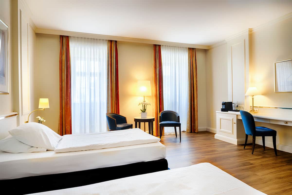 Ein Hotelzimmer mit zwei Betten, einem Schreibtisch, Stühlen, Lampen und Blick durch Vorhänge.
