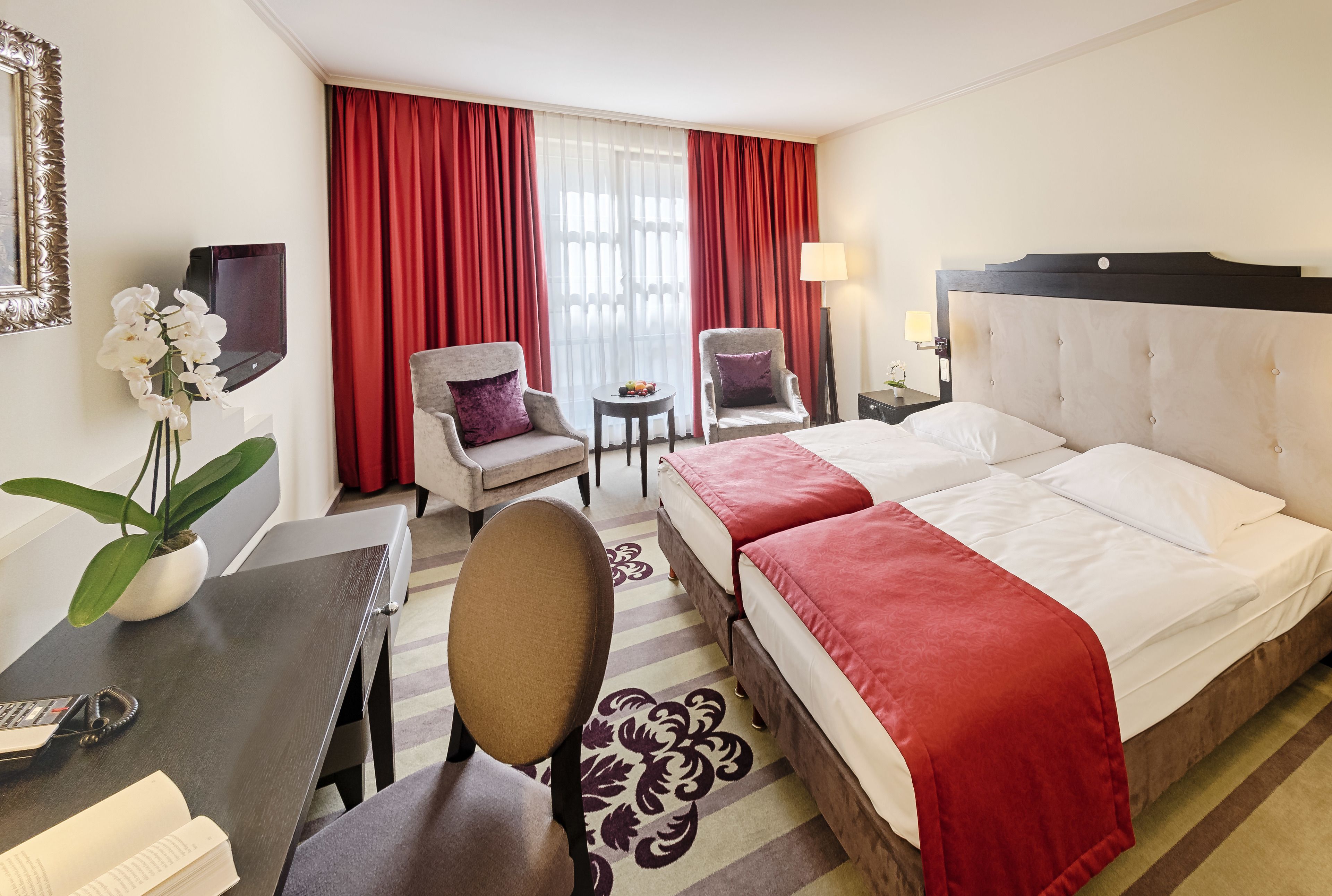 Habitación de hotel con cama, dos sillones, un escritorio con teléfono, una mesita con fruta, una planta en maceta y una ventana con cortinas rojas.