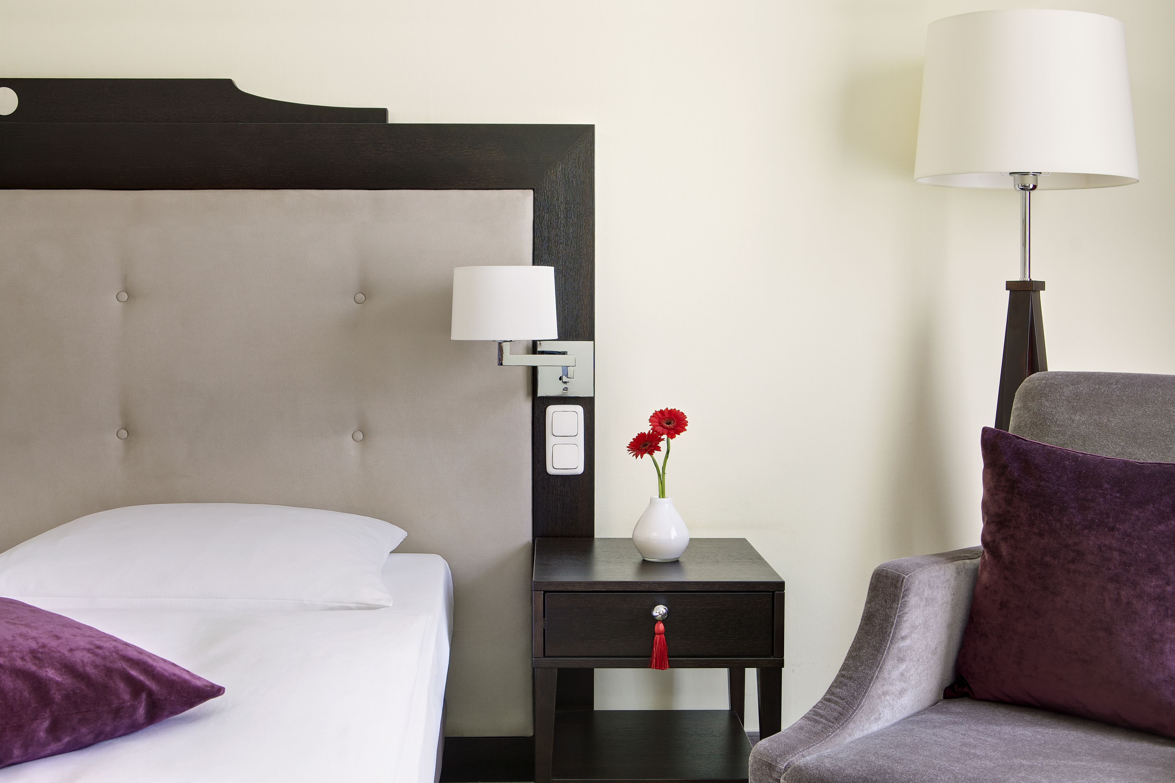 Una cama bien hecha con sábanas blancas, una mesita de noche con un jarrón blanco y flores rojas, una lámpara y un sillón gris con un cojín morado.