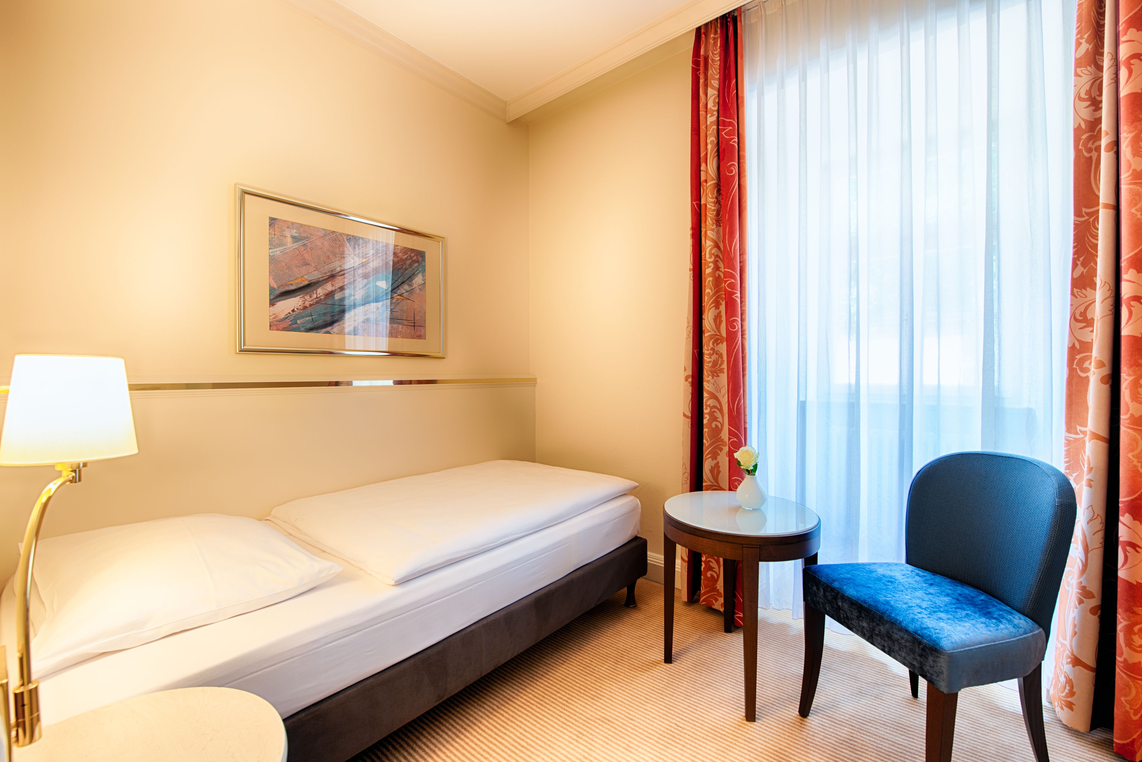 A hotel room with a bed, a chair, a small table, a lamp, and a painting on the wall.