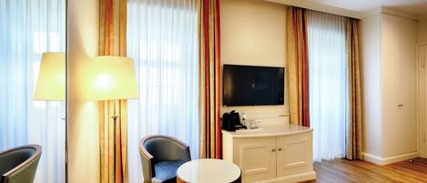 A hotel room with a TV, a small table, two chairs, a lamp, and a minibar.