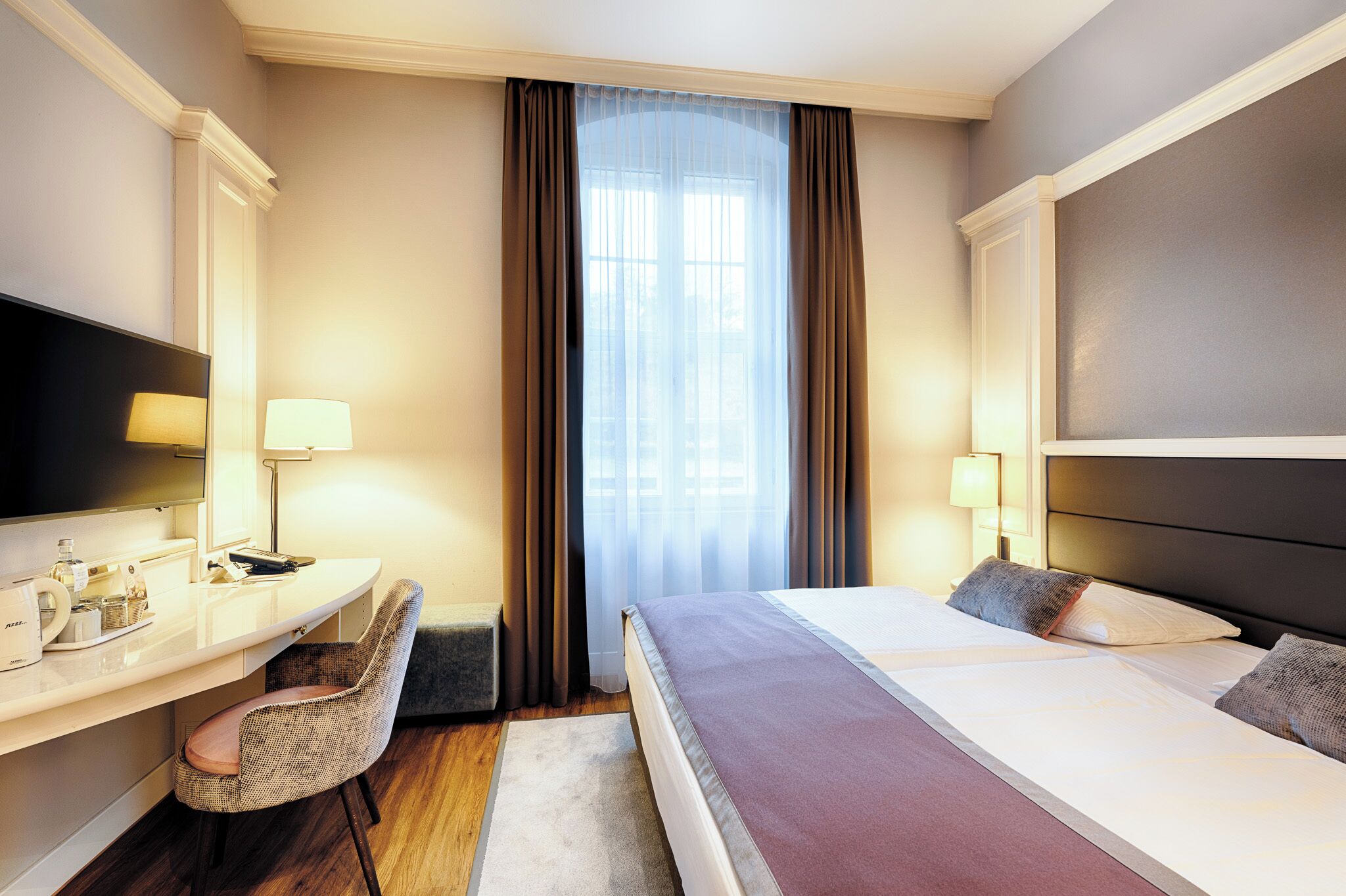 Una habitación de hotel con cama, escritorio con silla, televisor y ventana con cortinas.