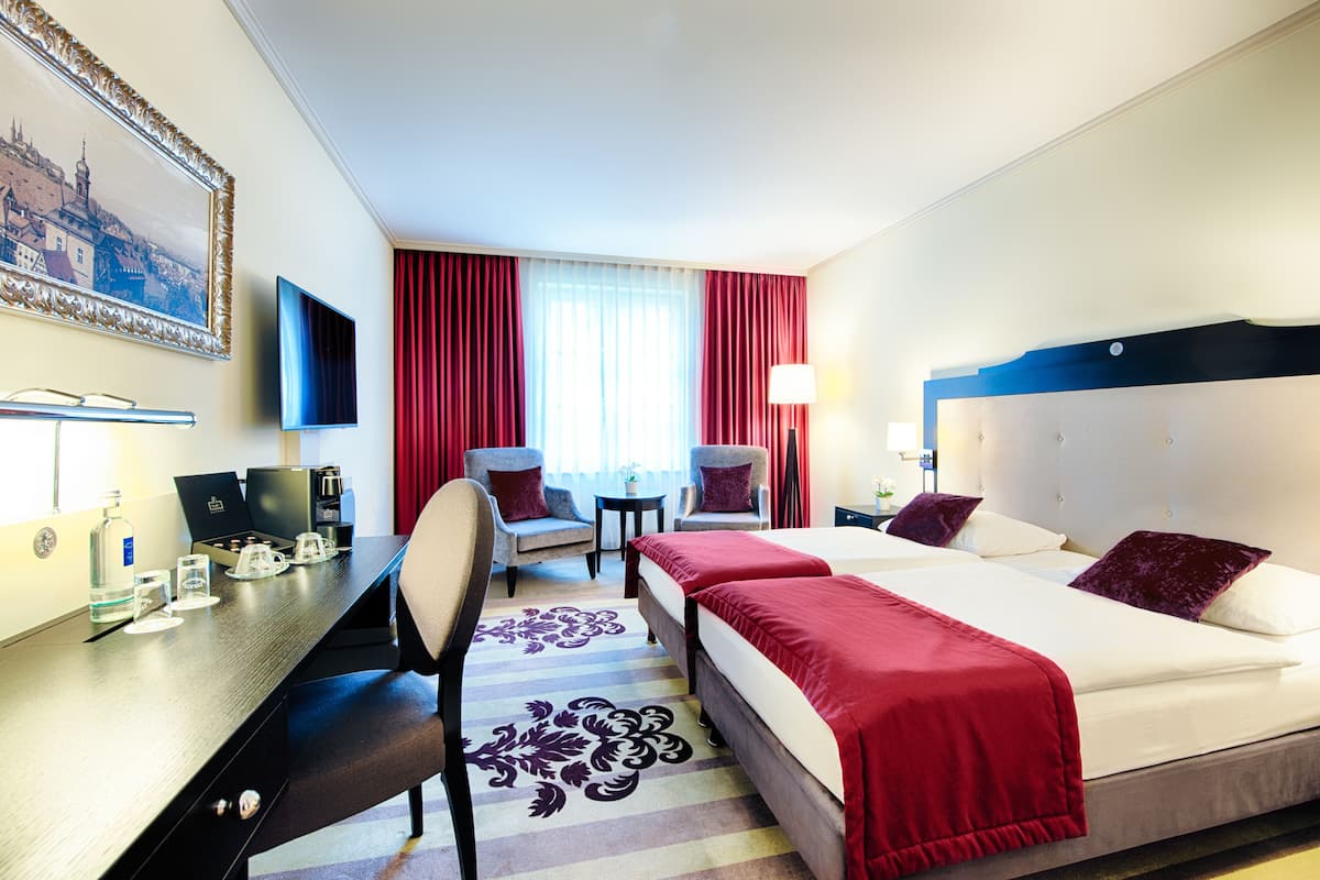 Ein Hotelzimmer mit einem großen Bett, zwei Sesseln, einem Schreibtisch mit Computer, einem Fernseher und einer Kaffeemaschine.
