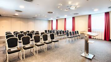A conference room with rows of chairs, a podium, and large windows with red curtains.