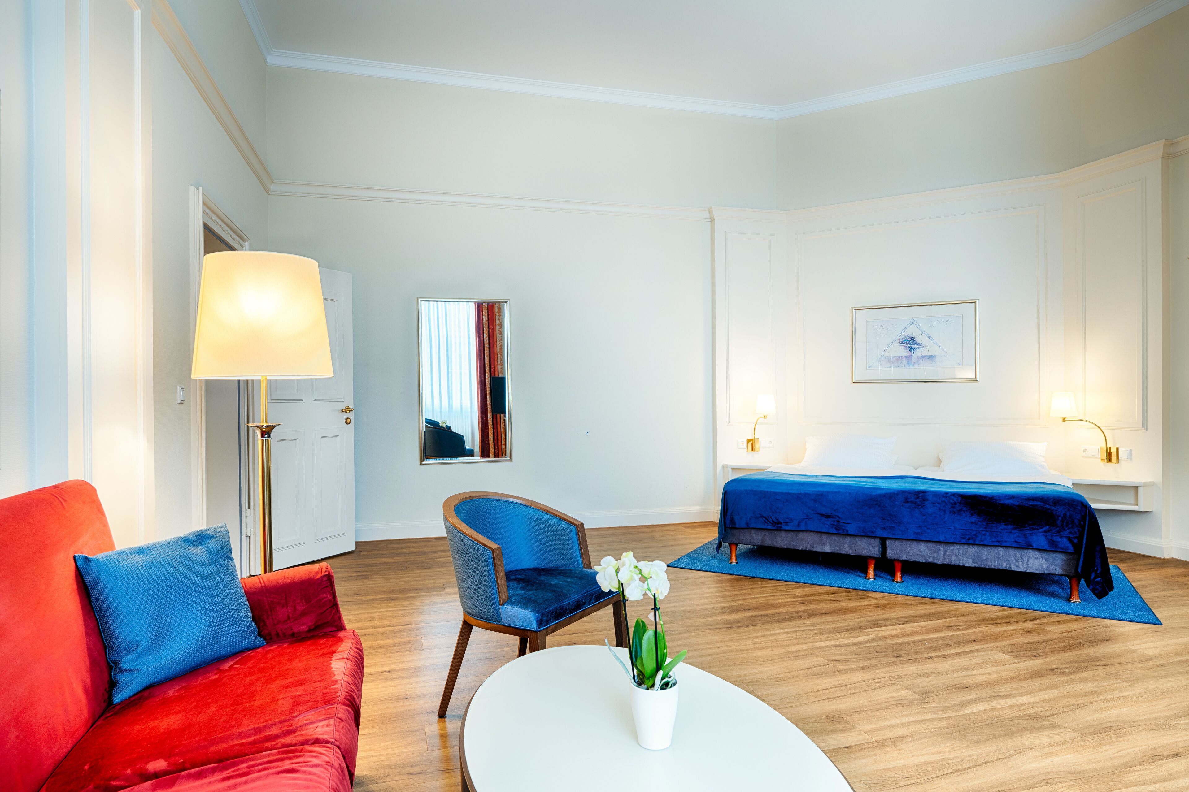 Ein Hotelzimmer mit rotem Sofa, blauem Sessel, weißem Couchtisch, Bett mit blauer Tagesdecke, einem Bild an der Wand und einer Lampe.